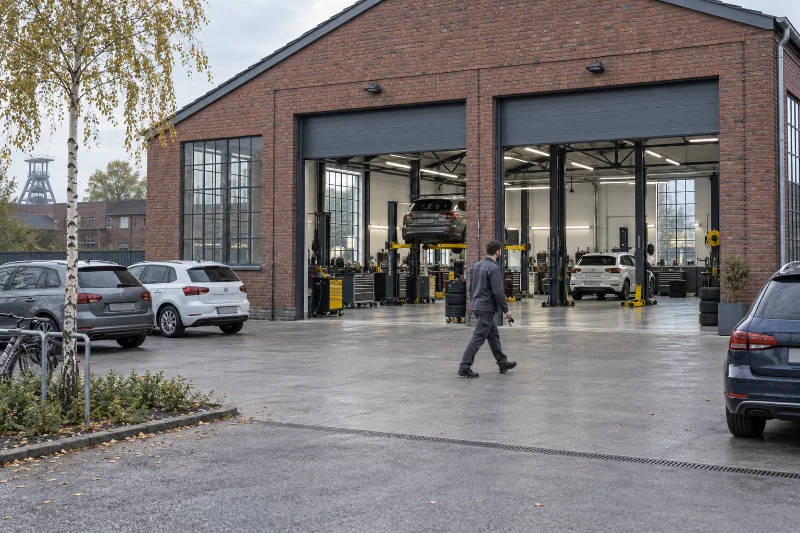 Backsteinhalle der Werkstatt mit zwei offenen Toren, davor der Hof mit parkenden Autos und einer Birke